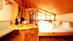Ein gemütliches Zimmer mit Holzboden, einem Bett mit weißer Bettwäsche, einem runden Holztisch mit Flasche und Glas sowie einem gemusterten Sofa.