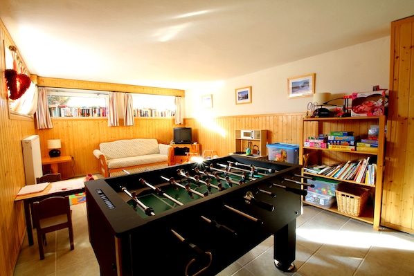 Una habitación acogedora con mesa de fútbol, sofá, televisión y estantería llena de libros y juegos.