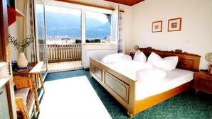 Ein Schlafzimmer mit einem großen Bett, einem Holzkopfende, einem Nachttisch, einer Kommode, einem Fenster mit Bergblick und einem Balkon.