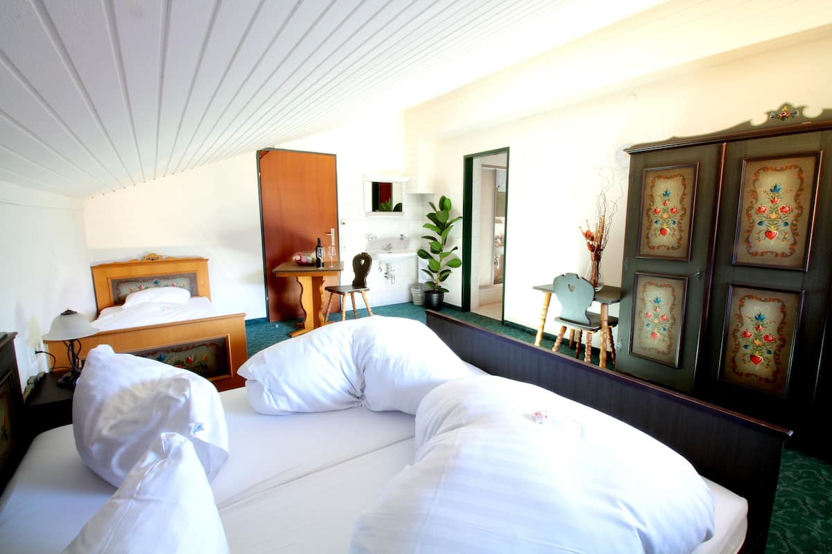 A hotel room with a neatly made bed, a wooden nightstand with a lamp, a small table with a chair, a mirror, and a cabinet with decorative panels.