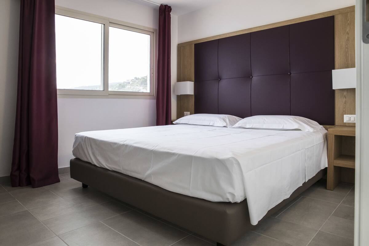 A modern hotel room with a large bed, a wooden headboard, two bedside lamps, and a window with curtains.