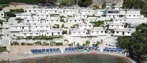 A coastal hotel with multiple white buildings, clear blue water, and a rocky hillside in the background.