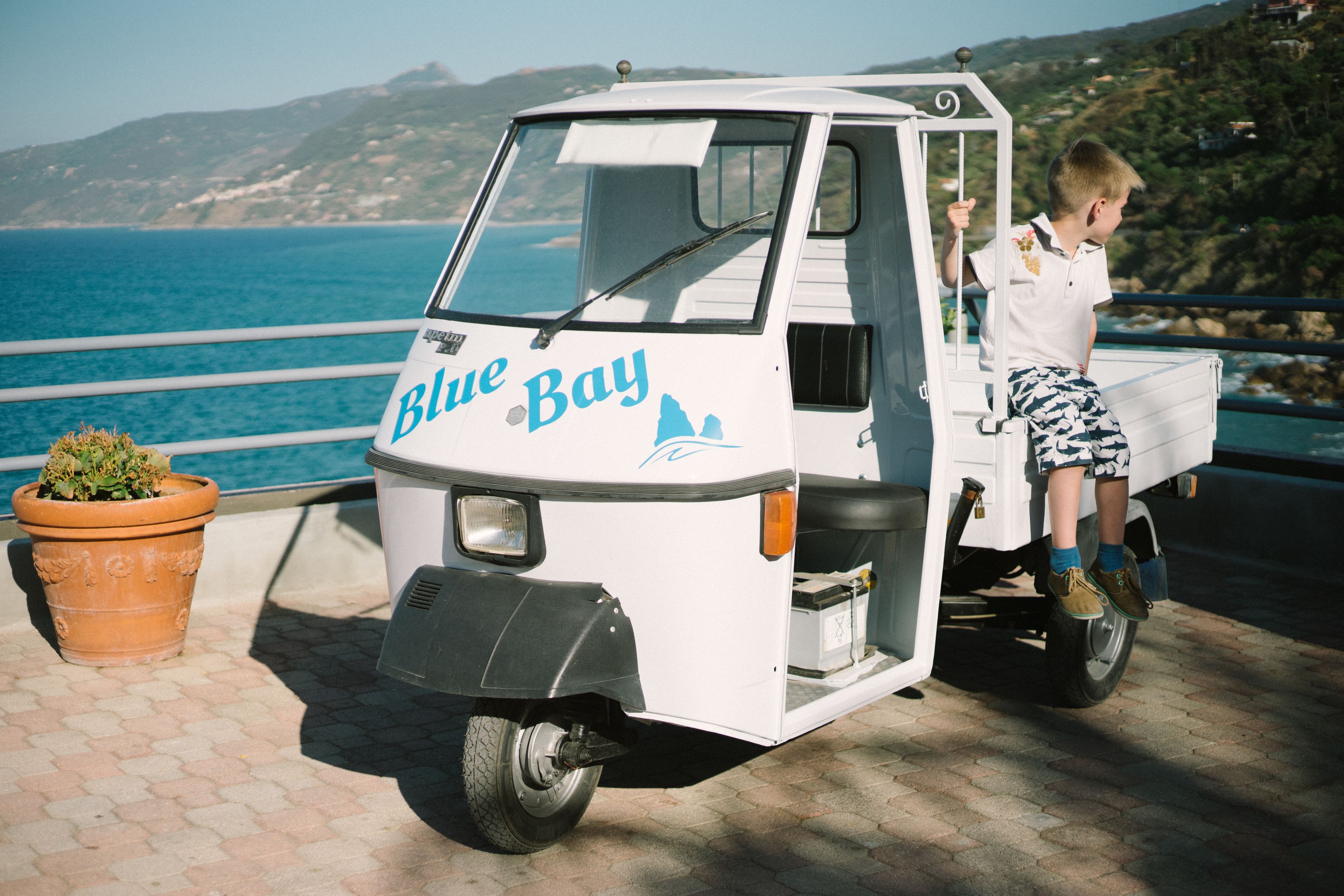 Un vehículo blanco pequeño con la leyenda "Blue Bay" estacionado en un área pavimentada, con vista al mar y colinas al fondo.
