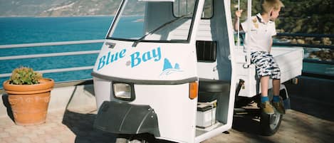 A small white vehicle with "Blue Bay" written on it, parked on a paved area with a view of the sea and hills in the background.