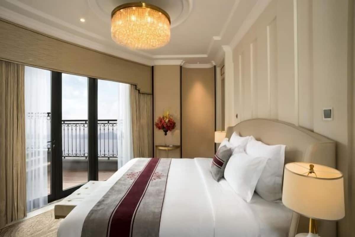 A hotel room with a large bed, a chandelier, a view of the outdoors, and a small table with a vase of flowers.