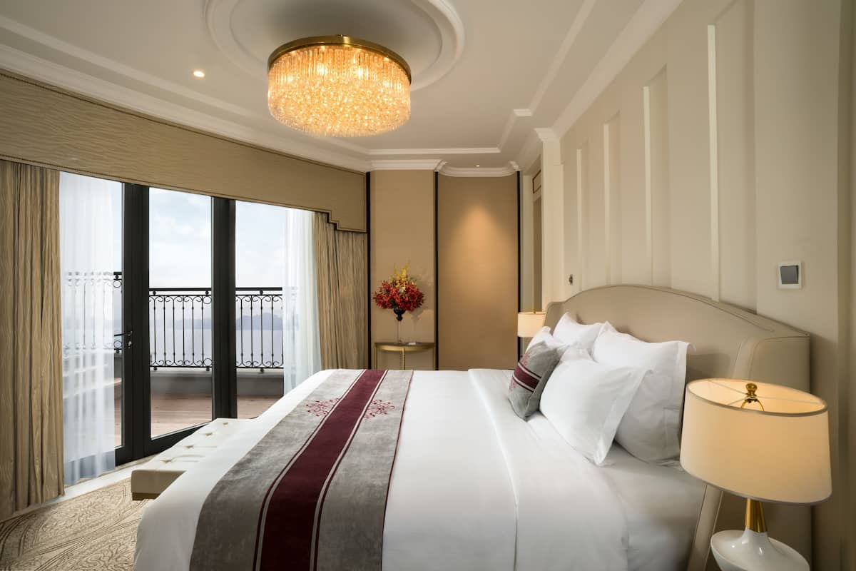 A hotel room with a large bed, a chandelier, a view of the city, and a balcony.