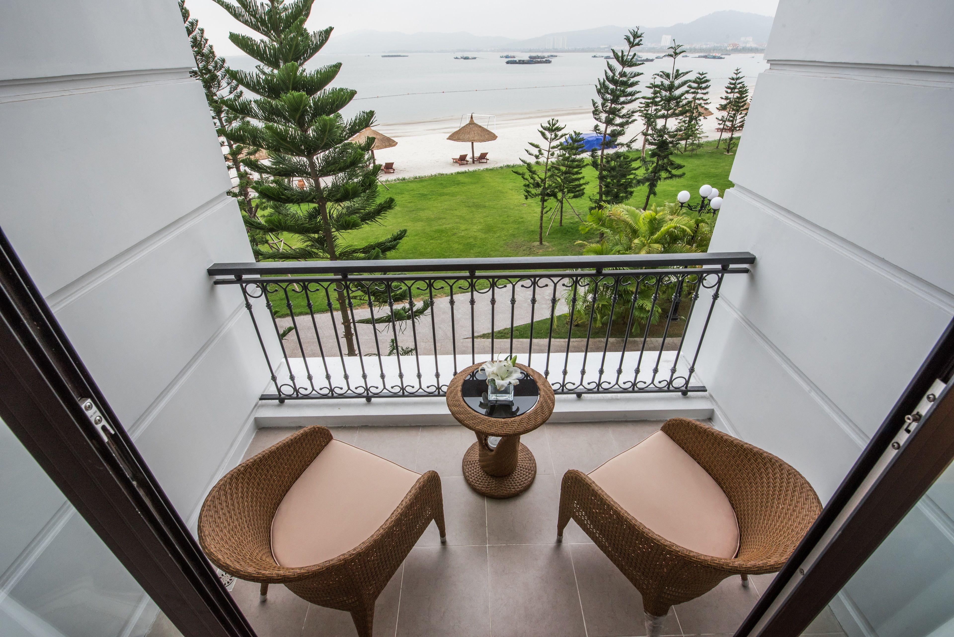 A balcony with wicker chairs and a small table, overlooking a beach with umbrellas and a clear view of the sea.