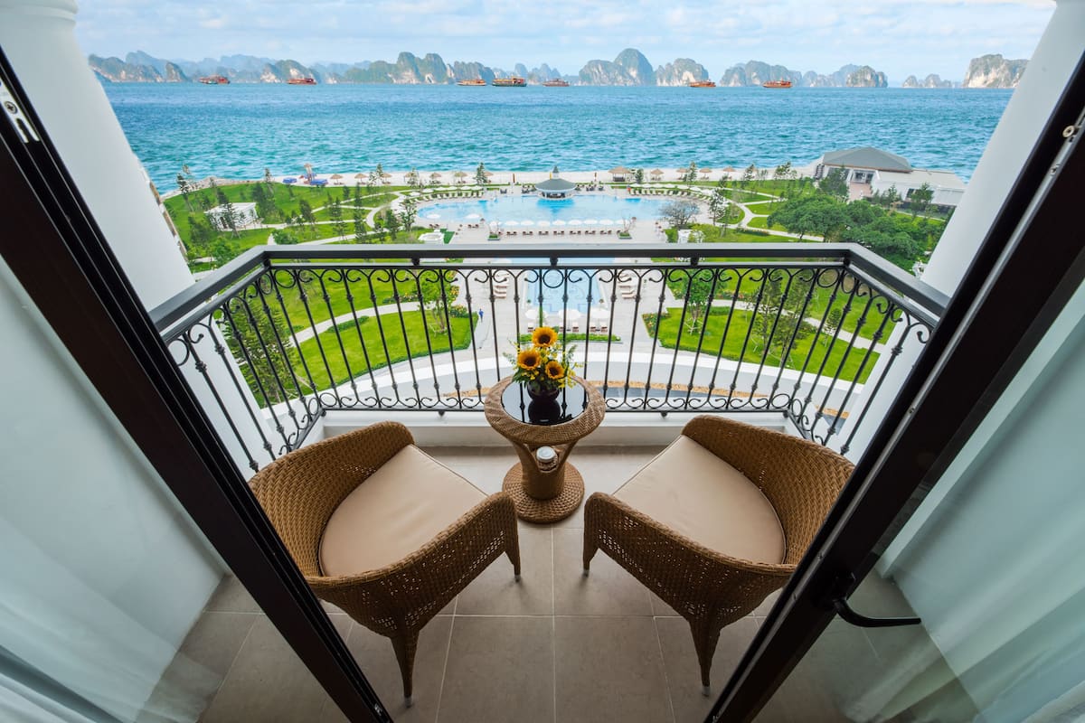 A balcony with wicker chairs and a table with a vase of flowers, overlooking a pool and a coastal view.