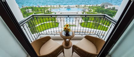 A balcony with wicker chairs and a table with a vase of flowers, overlooking a pool and a coastal view.