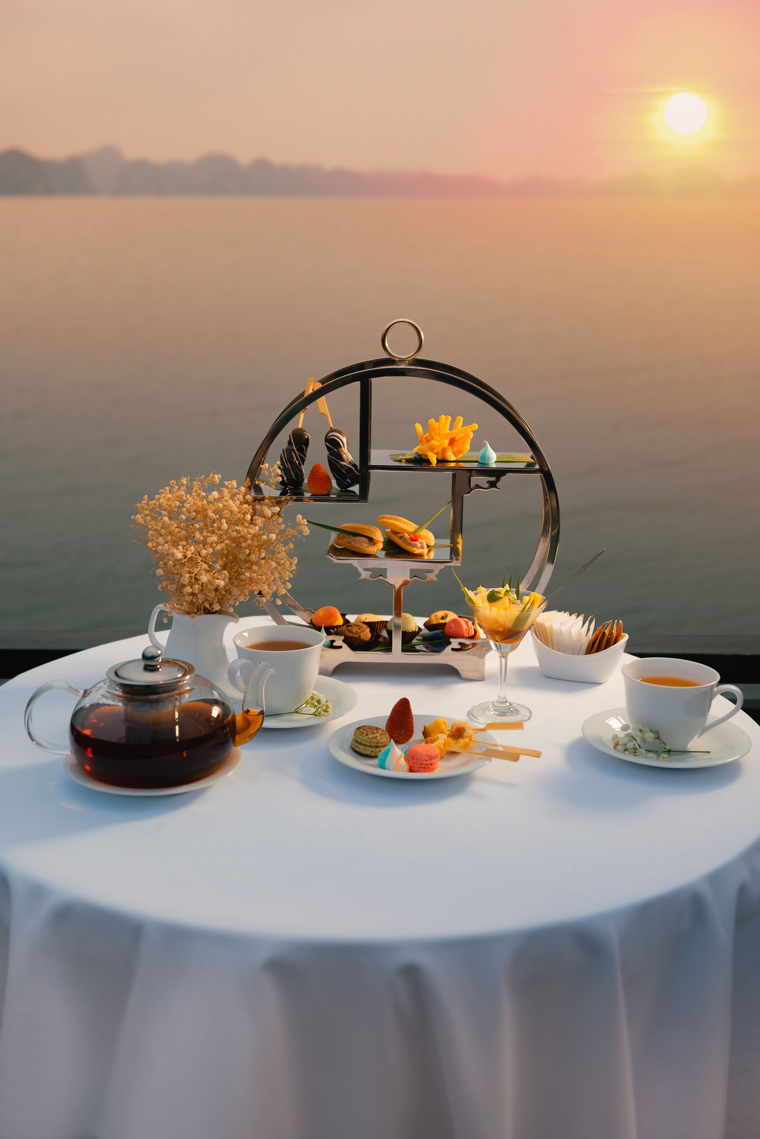 Una mesa dispuesta con té, pasteles y una tetera, con vista a un cuerpo de agua al atardecer.