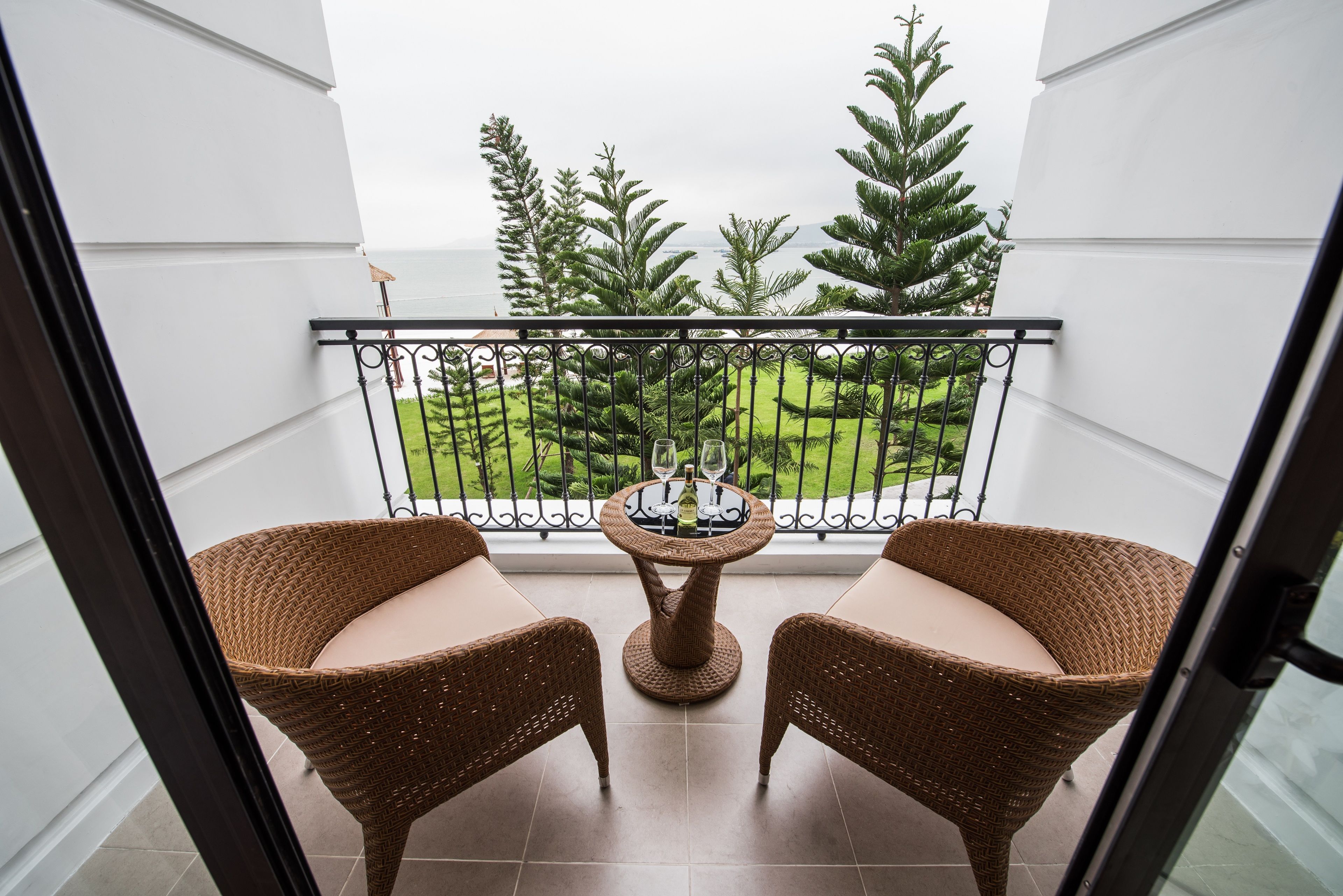 Un balcón con sillas de mimbre y una pequeña mesa, con vistas a un jardín cuidado y una masa de agua.