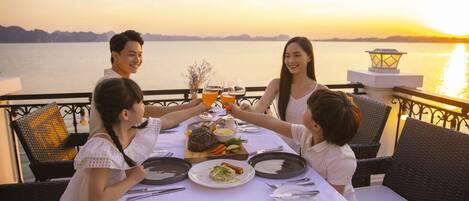 A table set for four people with a meal and drinks, overlooking a body of water at sunset.