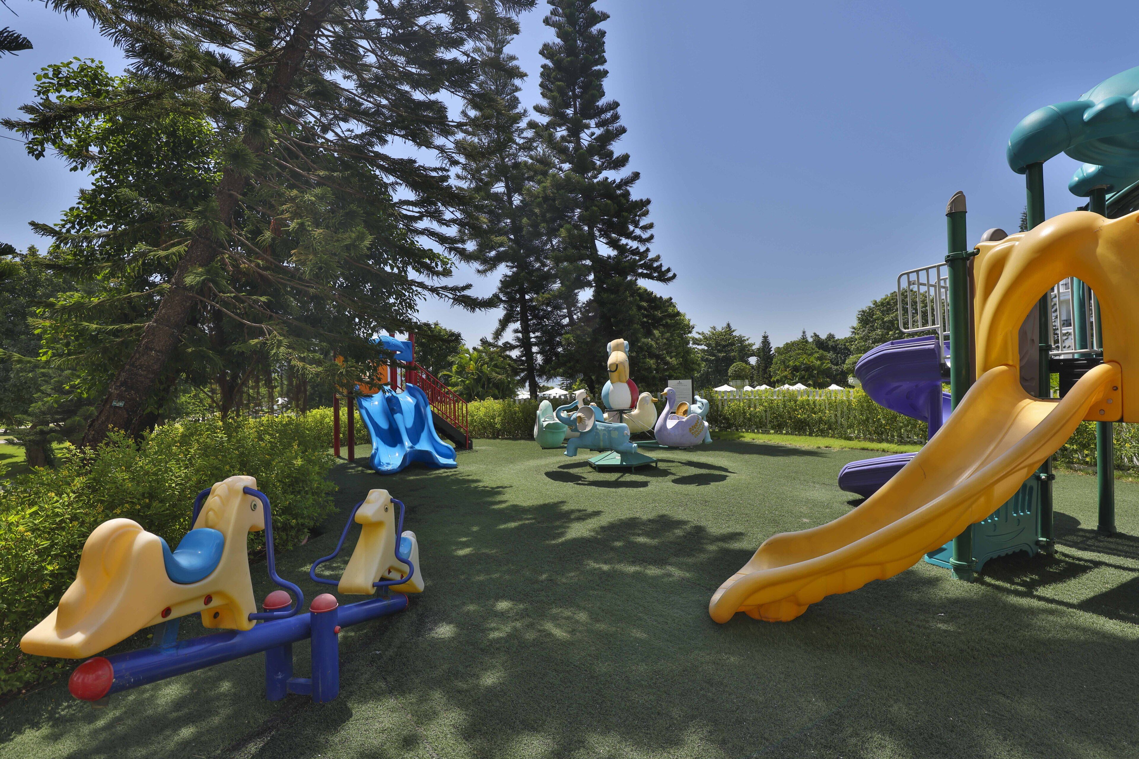A playground with various play structures, including slides and swings, surrounded by trees and greenery.