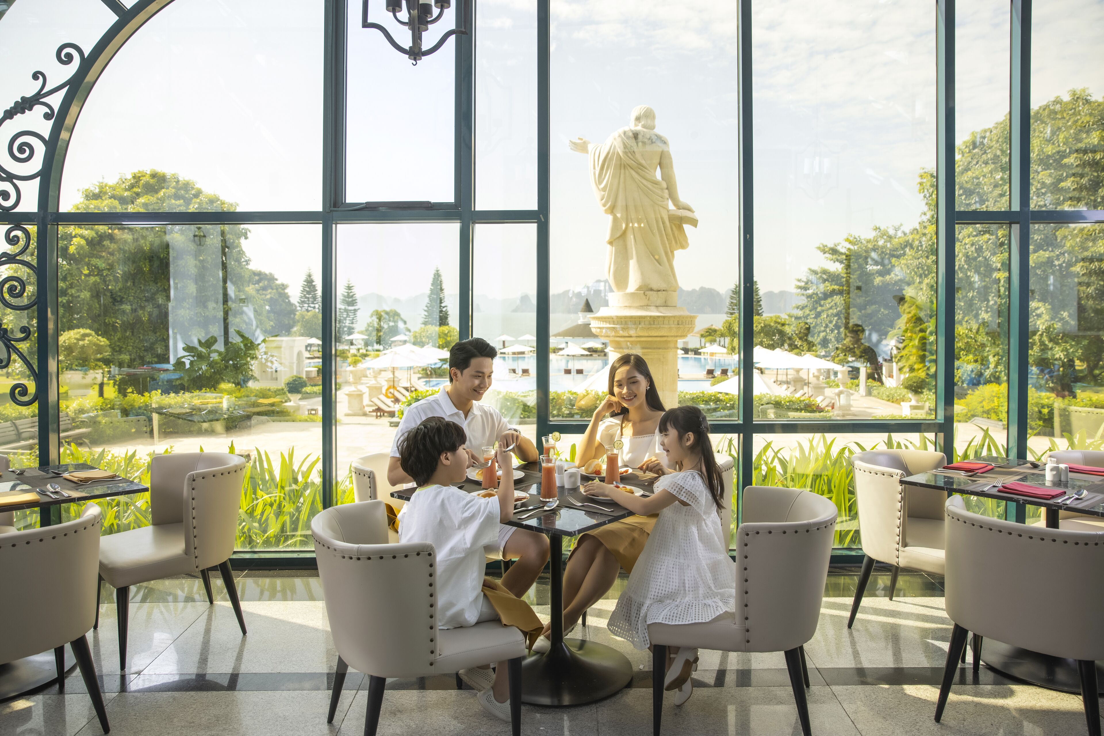 A group of people dining at a table with a view of a statue and greenery outside.