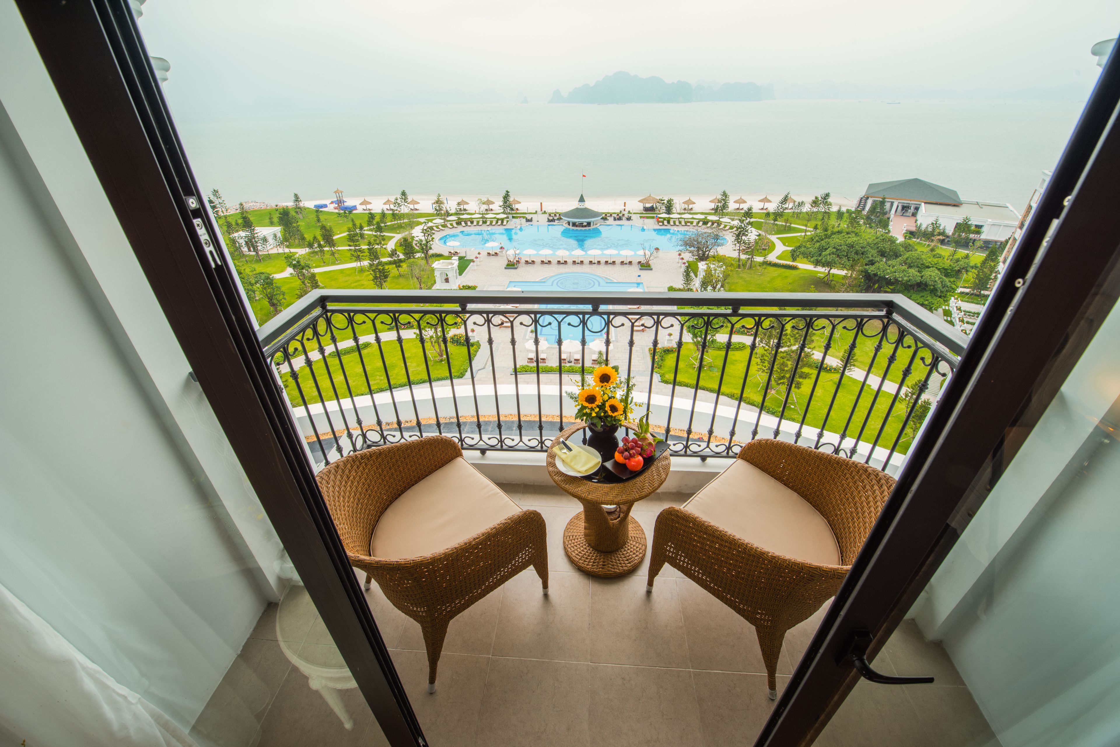 A balcony with wicker chairs, a small table with a vase of flowers, and a view of a swimming pool and beach.