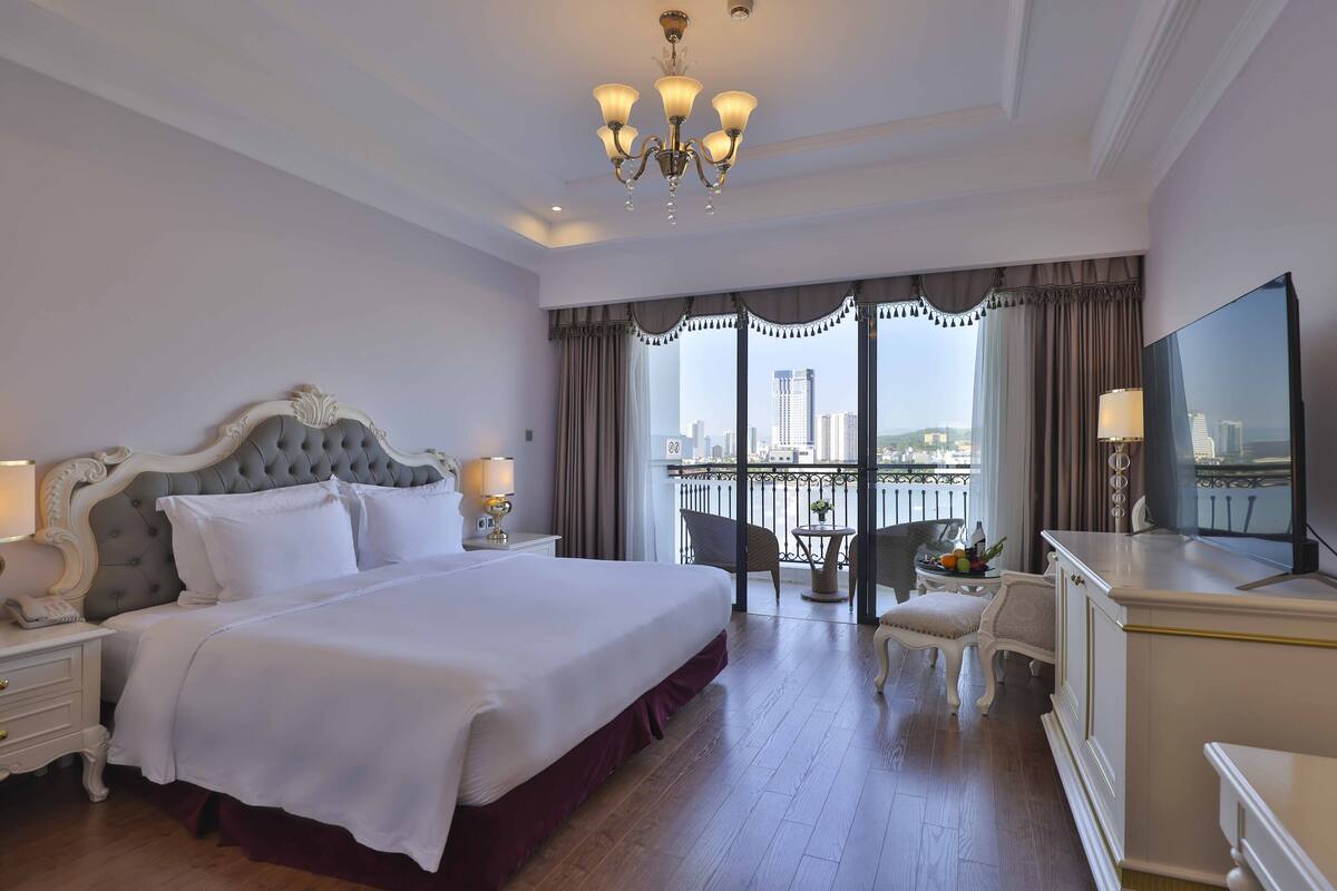 A hotel room with a large bed, a TV, a desk, and a balcony with a city view.