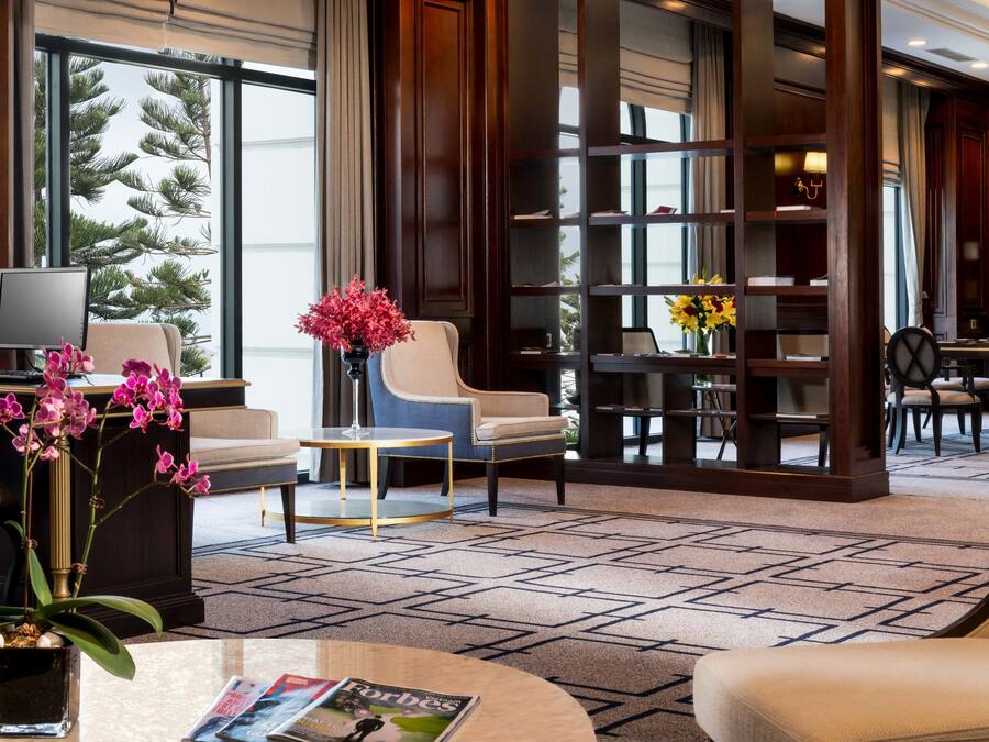 A hotel lobby with a seating area, a coffee table, a bookshelf, and a large window with a view of trees.