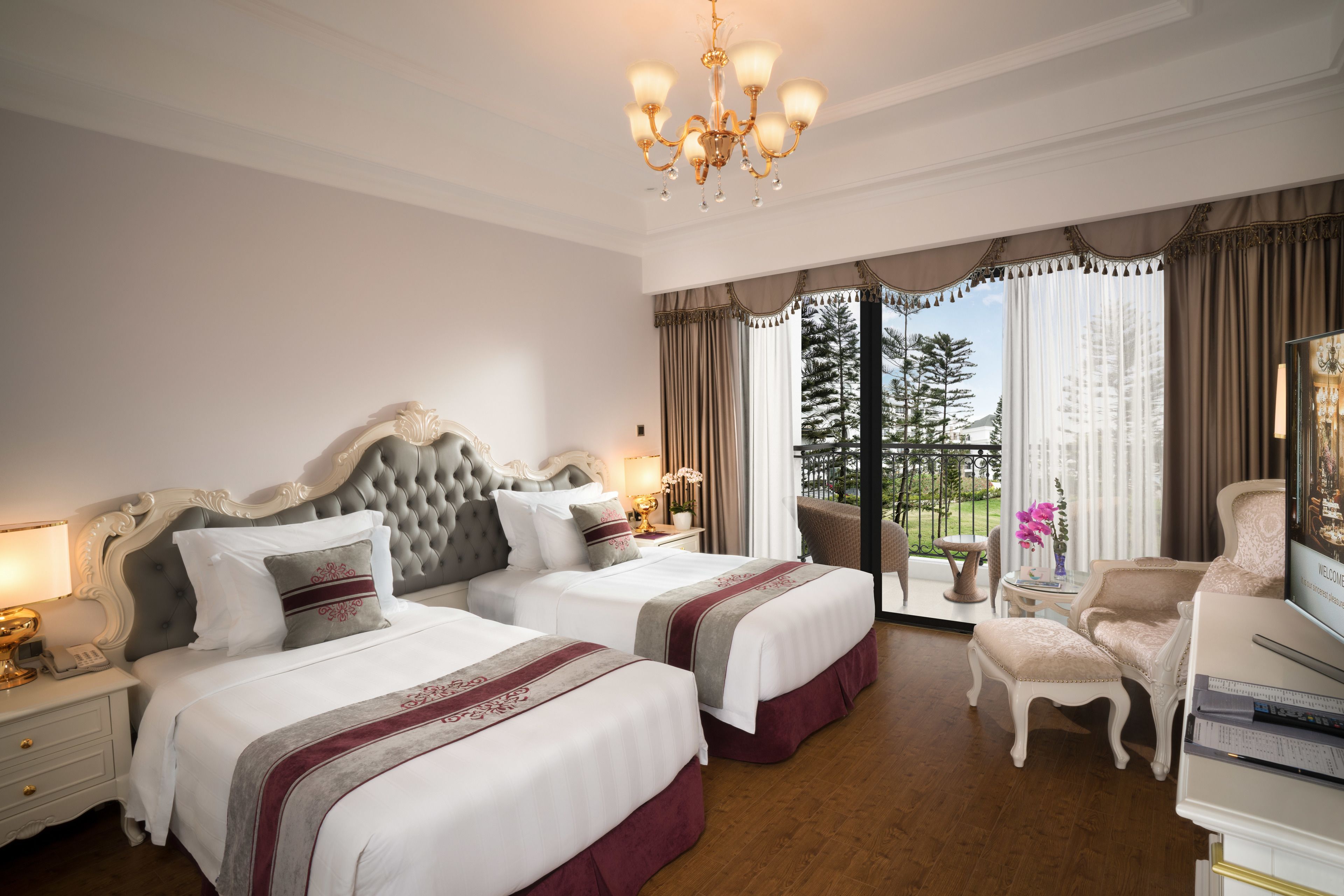 Une chambre d’hôtel avec deux lits, un lustre, un balcon donnant sur la verdure et un coin salon avec un fauteuil et une table.