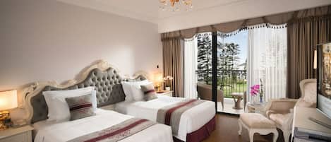 A hotel room with two beds, a chandelier, a balcony with a view of trees, and a sitting area with a chair and a table.