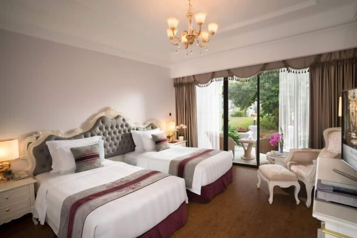 A hotel room with two beds, a chandelier, a sitting area with a chair and table, and a view of the outdoors.