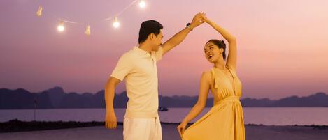 Una pareja bailando en la playa al atardecer, con luces colgantes por encima.