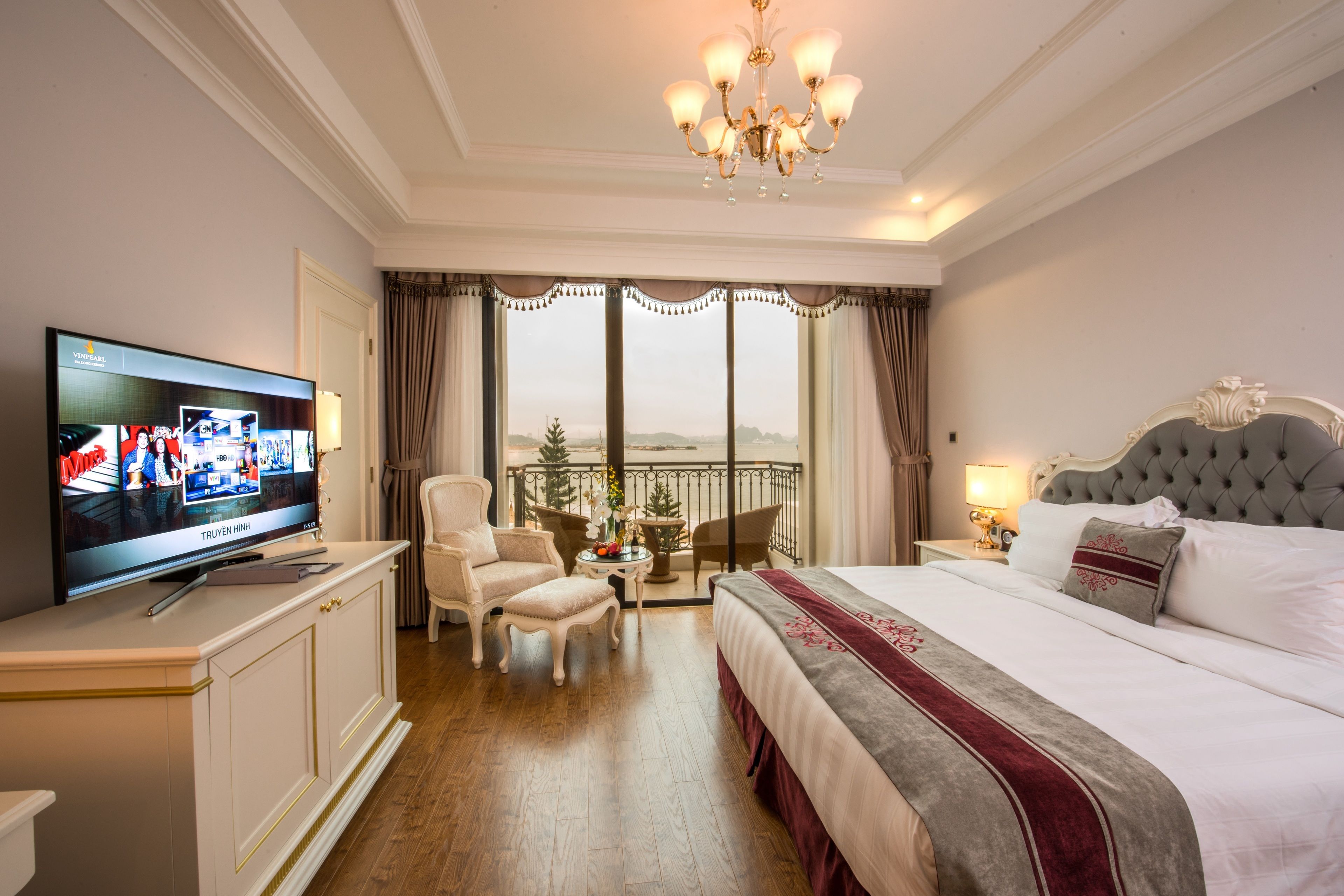 Habitación de hotel con un televisor de pantalla plana grande, cama con cabecera tapizada en gris, candelabro, balcón con vistas y una pequeña mesa con sillas.