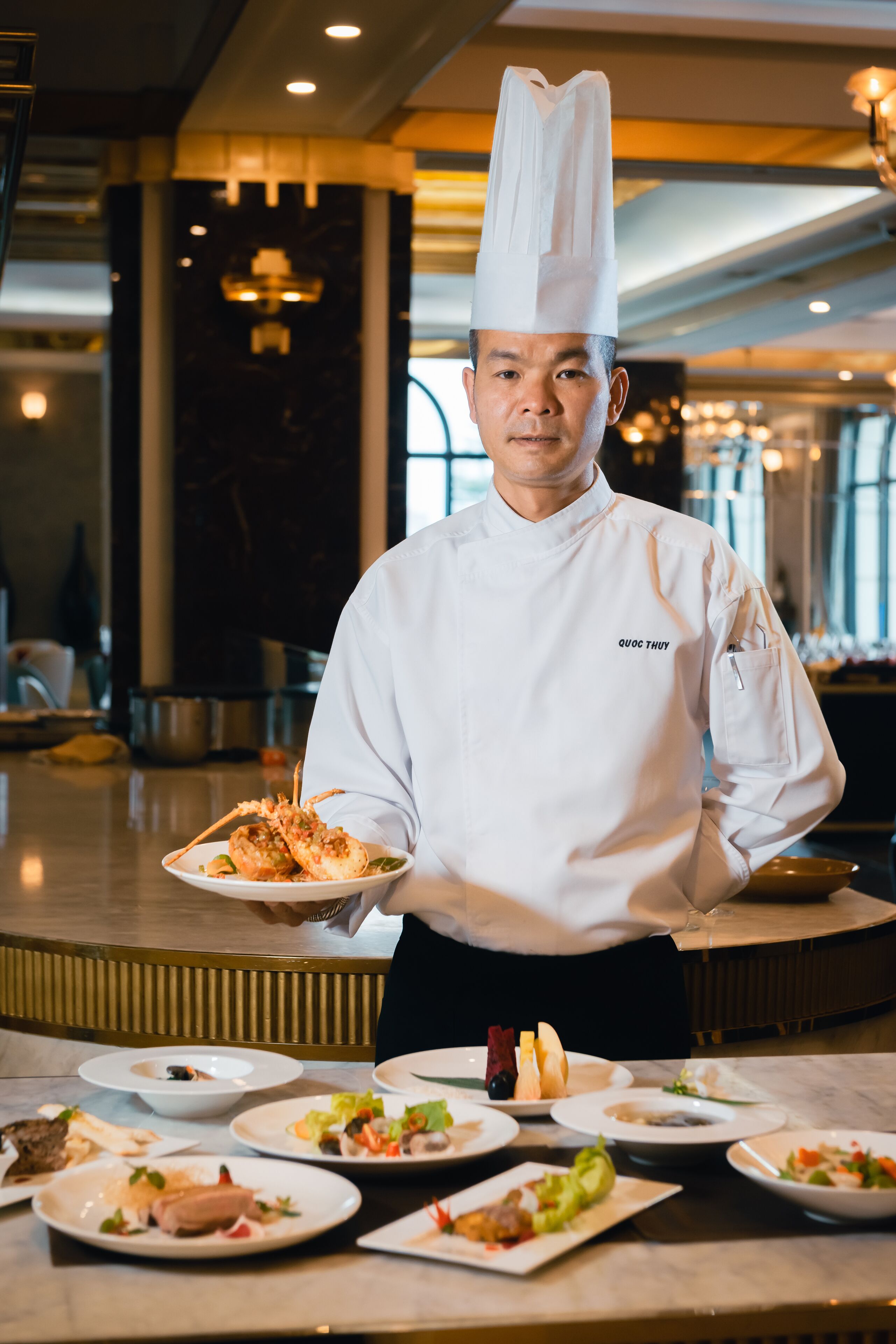Un chef, vêtu d’un uniforme blanc et coiffé d’un haut-de-forme, tient une assiette de nourriture dans un espace de restauration moderne et bien éclairé, où plusieurs plats sont disposés sur la table.