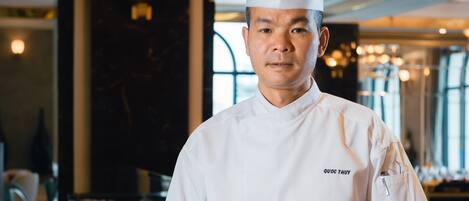 Un chef, vêtu d’un uniforme blanc et coiffé d’un haut-de-forme, tient une assiette de nourriture dans un espace de restauration moderne et bien éclairé, où plusieurs plats sont disposés sur la table.