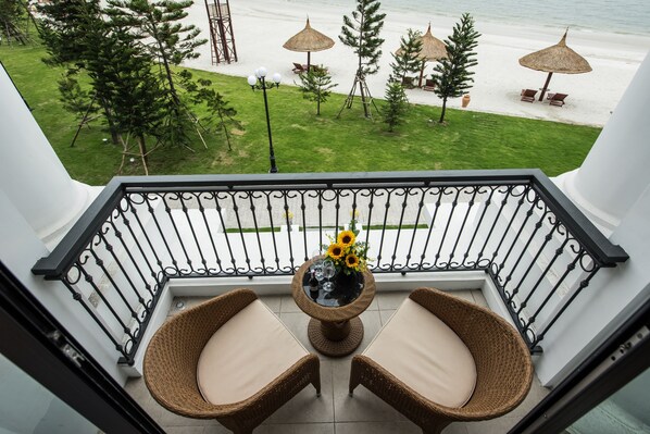 A balcony with wicker chairs and a table with a vase of sunflowers, overlooking a beach with umbrellas and lounge chairs. - Vinpearl Resort & Spa Ha Long (Bai Chay)