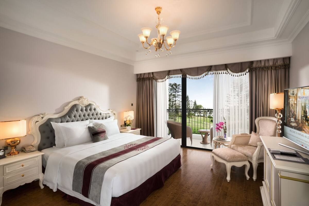A spacious bedroom with a large bed, a chandelier, a desk with a computer, and a view of the outdoors.