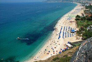 Una playa costera con hileras de sombrillas y sillas, un mar azul y cristalino, y un acantilado rocoso que domina la playa.