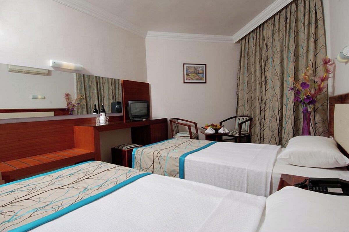 A hotel room with two beds, a desk with a television, a chair, and a small table with a vase of flowers.