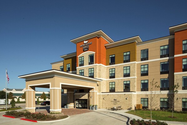 Un hotel de varios pisos con un llamativo letrero de "Homewood Suites", un acceso cubierto y un área frontal ajardinada.