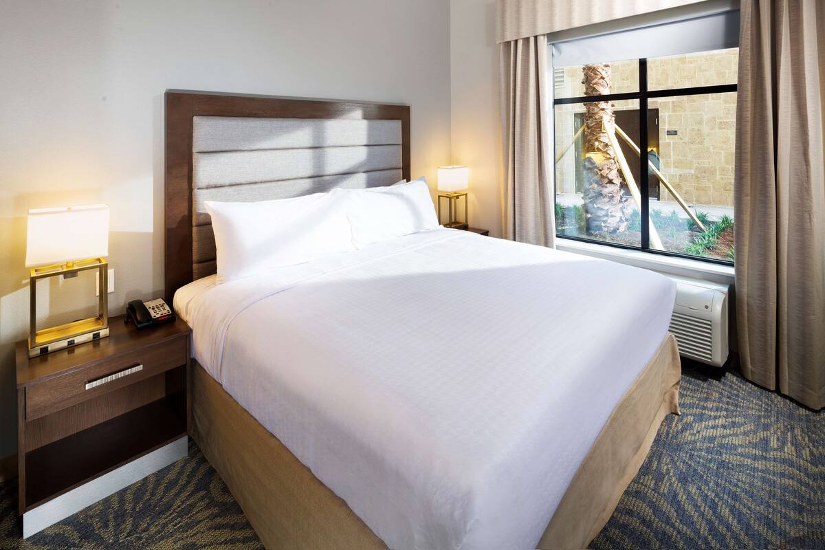 Habitación de hotel con una cama grande, lámparas de noche, teléfono y ventana con cortinas.