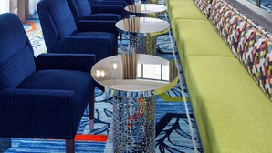 Un hall d’hôtel moderne avec un long canapé, des fauteuils bleus et un divan vert. On y trouve des tables rondes et un tapis à motifs.