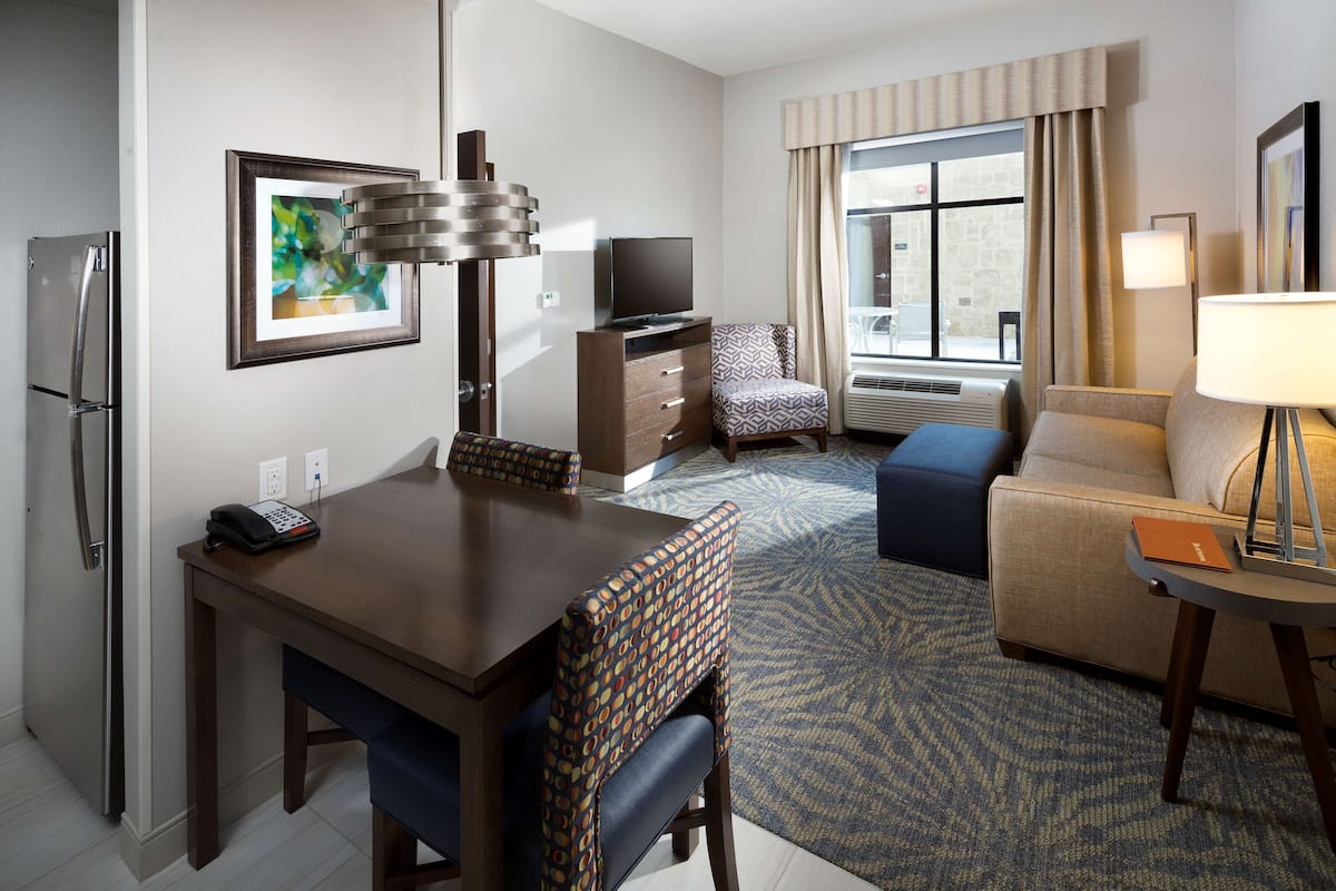 Habitación de hotel con una mesa de comedor, sillas, televisor, sofá, lámpara y ventana con cortinas.