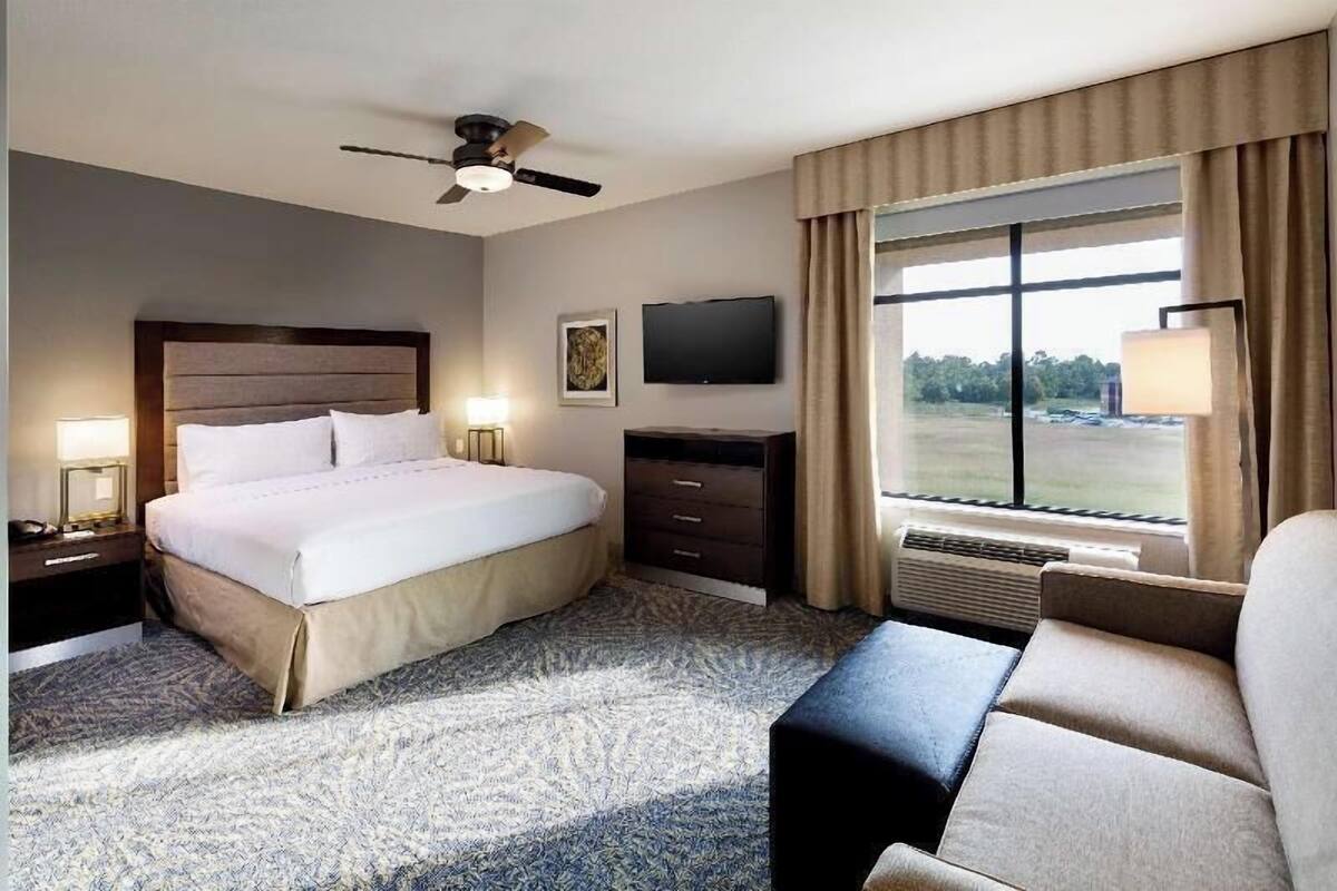Habitación de hotel con cama, televisión, ventilador de techo, ventana con cortinas y escritorio con lámpara.