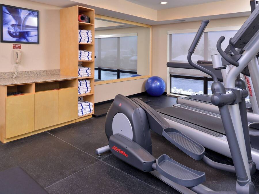 Un gimnasio con cintas de correr, una bicicleta estática y una pelota de ejercicios azul. Hay un mueble de madera con toallas y un teléfono en la pared.