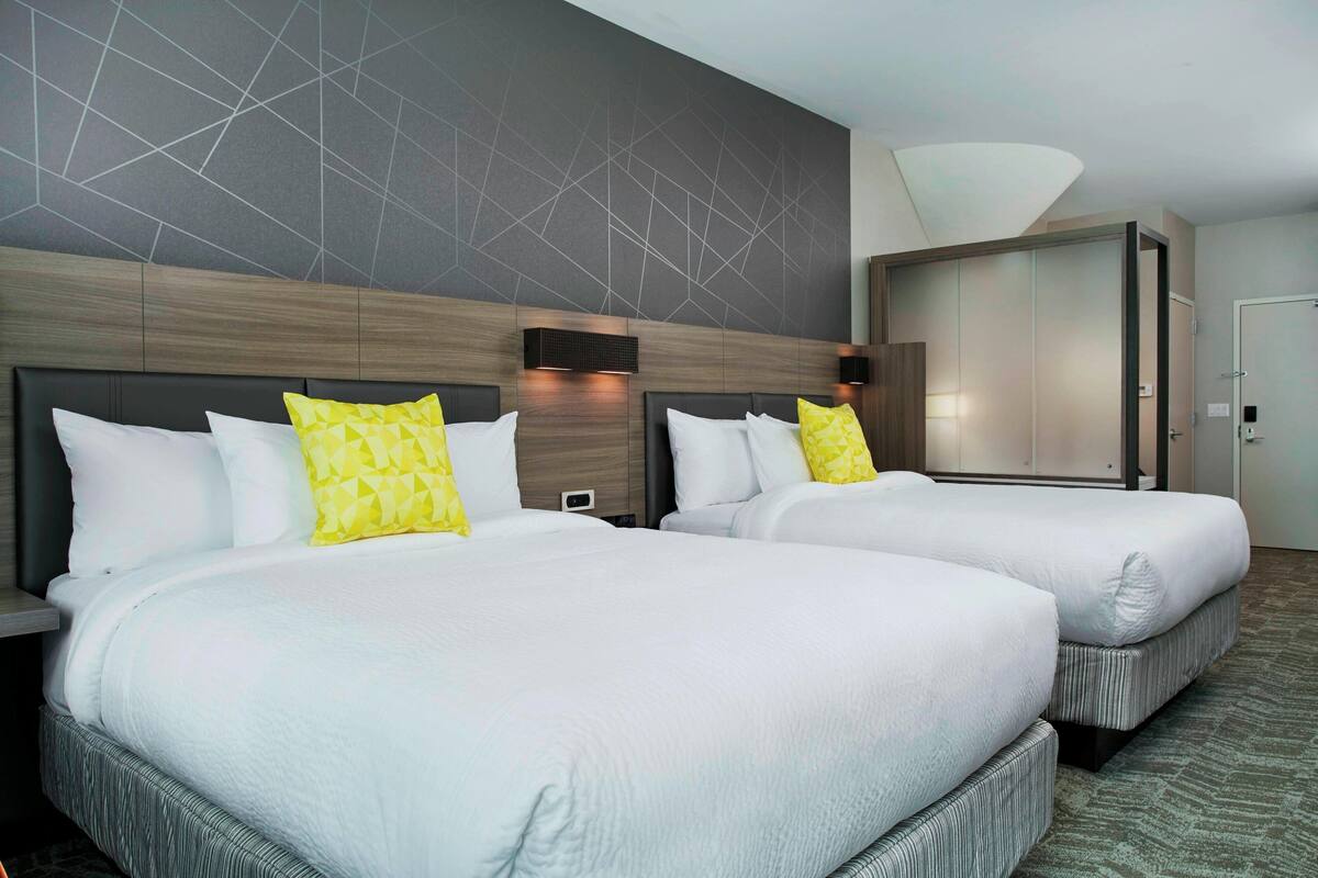 A hotel room with two beds, each with white bedding and a single yellow pillow. The room features a modern design with a geometric-patterned wall and a large, white ceiling light.
