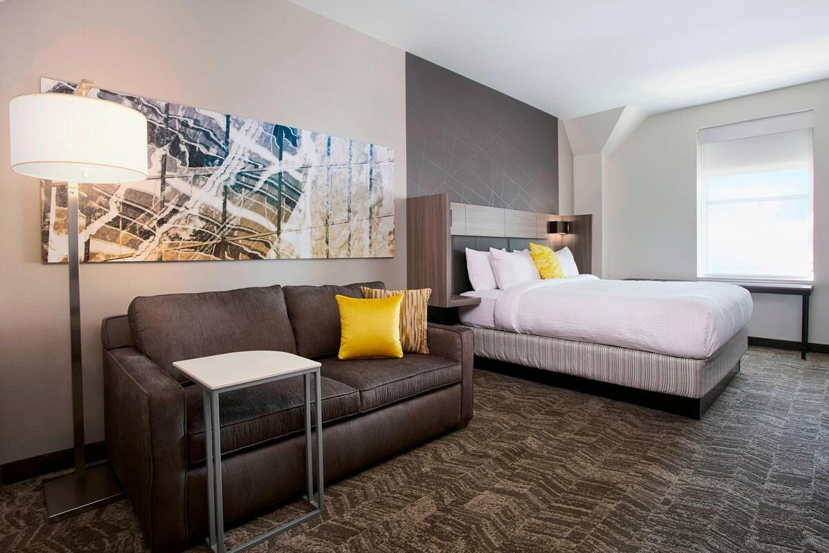 A hotel room with a brown leather sofa, a small table, a bed with white bedding, a large abstract painting, and a window with a view.