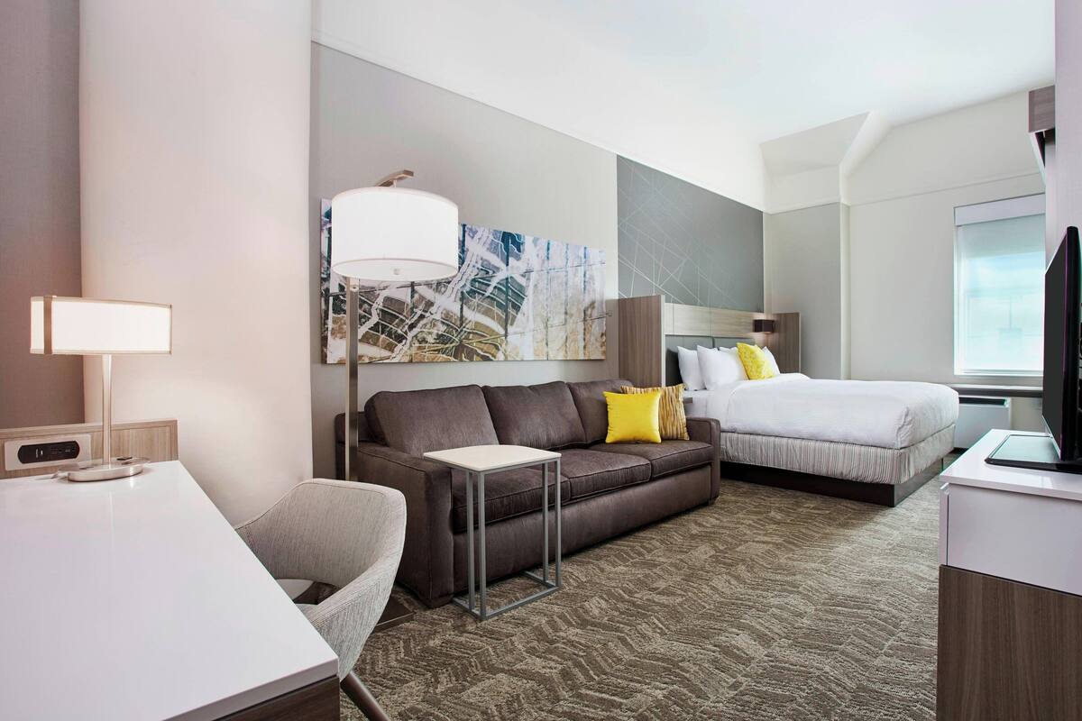A modern hotel room with a sofa, a small table, a bed with pillows, a TV, and a large window.
