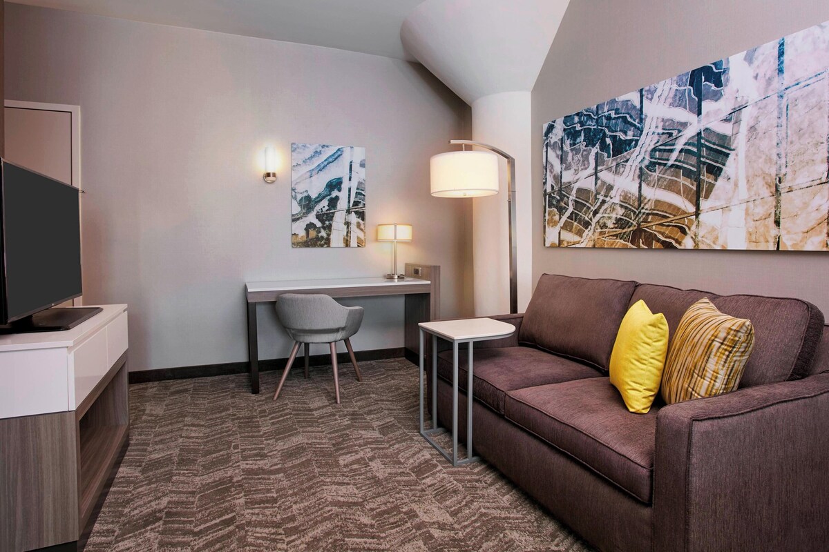 A hotel room with a brown sofa, a small desk with a chair, a television, and two wall-mounted lamps.