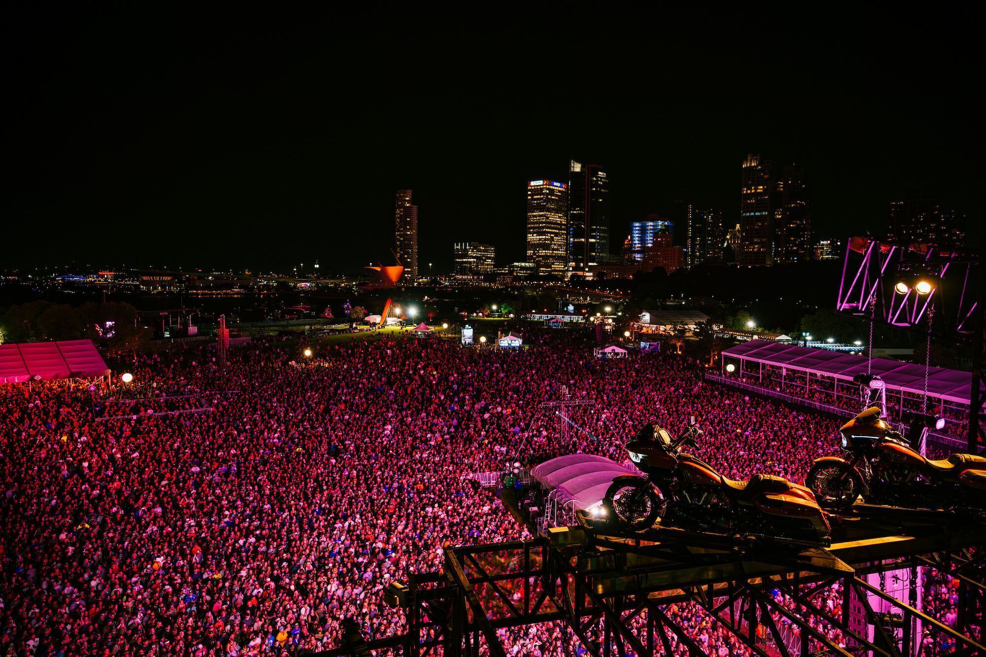 A large outdoor concert with a crowd, a stage, and a city skyline in the background.