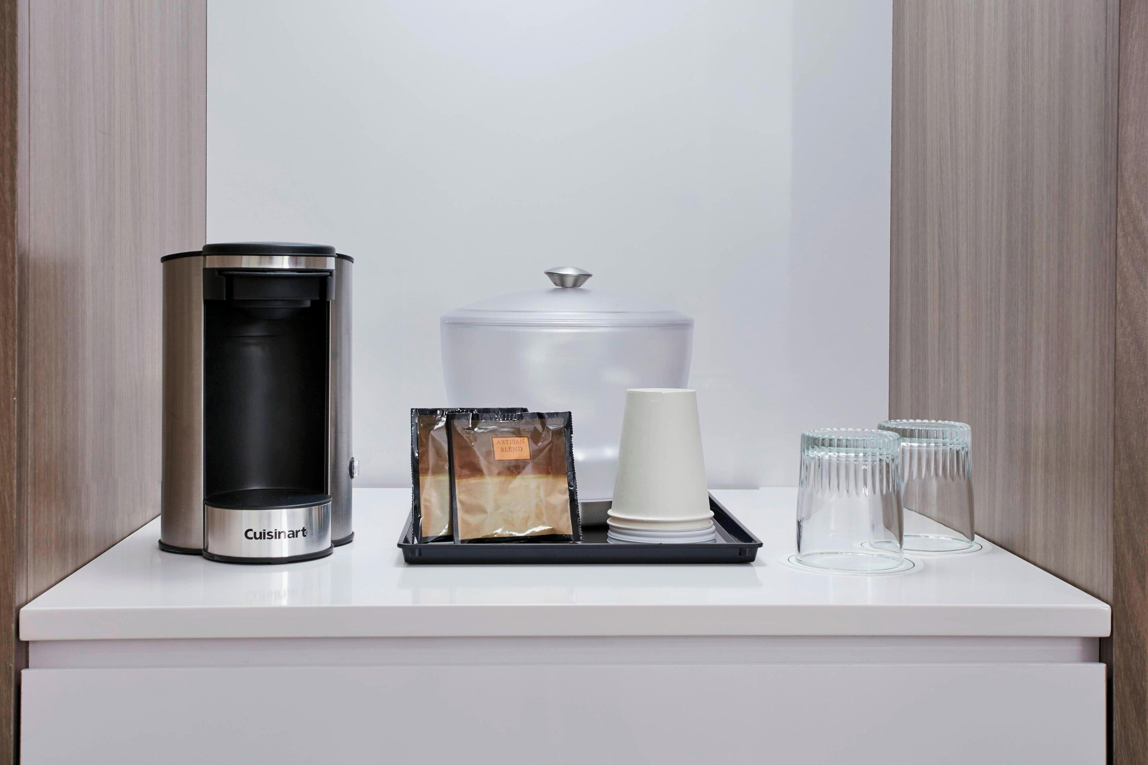 Une machine à café, une cafetière, un plateau avec un sachet de café, une tasse blanche et deux verres sur un comptoir blanc.