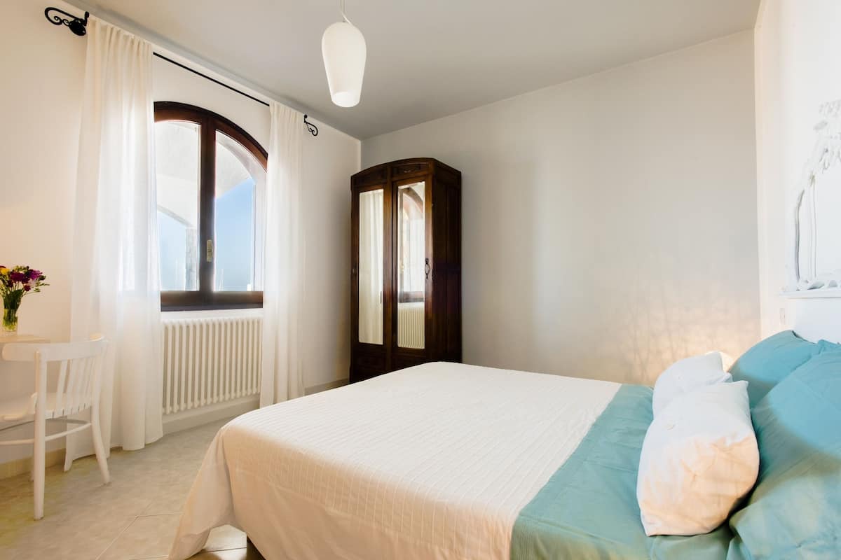 Un dormitorio con cama, un armario de madera, una ventana con cortinas, un radiador y una pequeña mesa con silla.