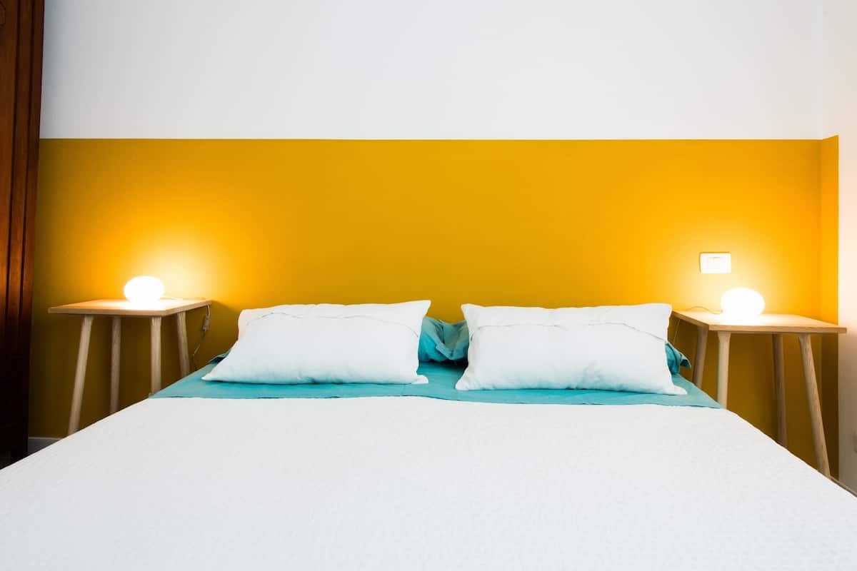 Una cama con ropa de cama blanca, dos almohadas blancas y una colcha azul verdosa. Hay dos mesitas de noche con lámparas.