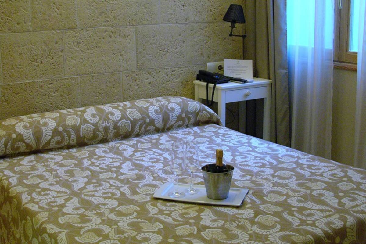 Een hotelkamer met een bed met een patroon, een nachtkastje met een lamp, een telefoon en een fles champagne.