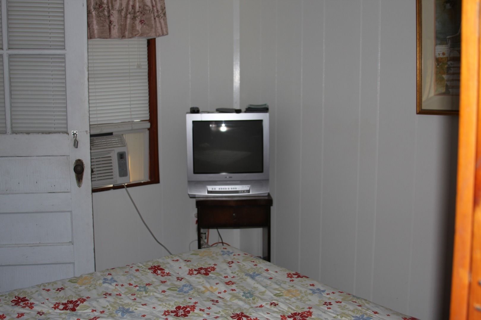 A bedroom with a bed, a television on a stand, a door, and a window with blinds.