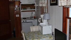 A compact kitchen with a stove, sink, and microwave. There is a dining table with a floral tablecloth and a chair. A lamp is on the table, and a picture hangs on the wall.