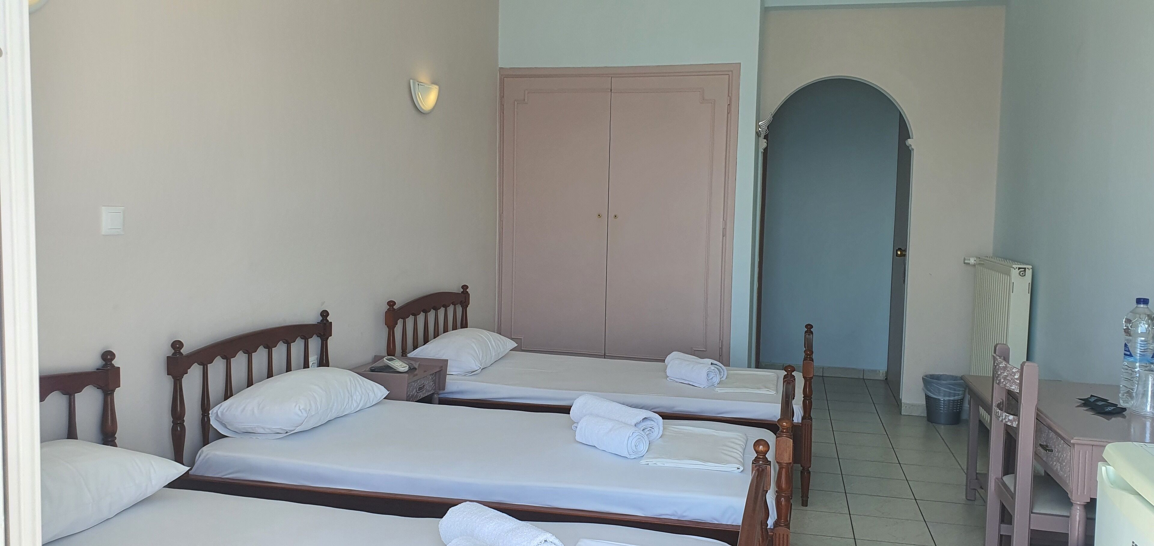 A hotel room with three single beds, a wooden headboard, a bedside table with a phone, a wall-mounted light, and a large arched doorway leading to another room.